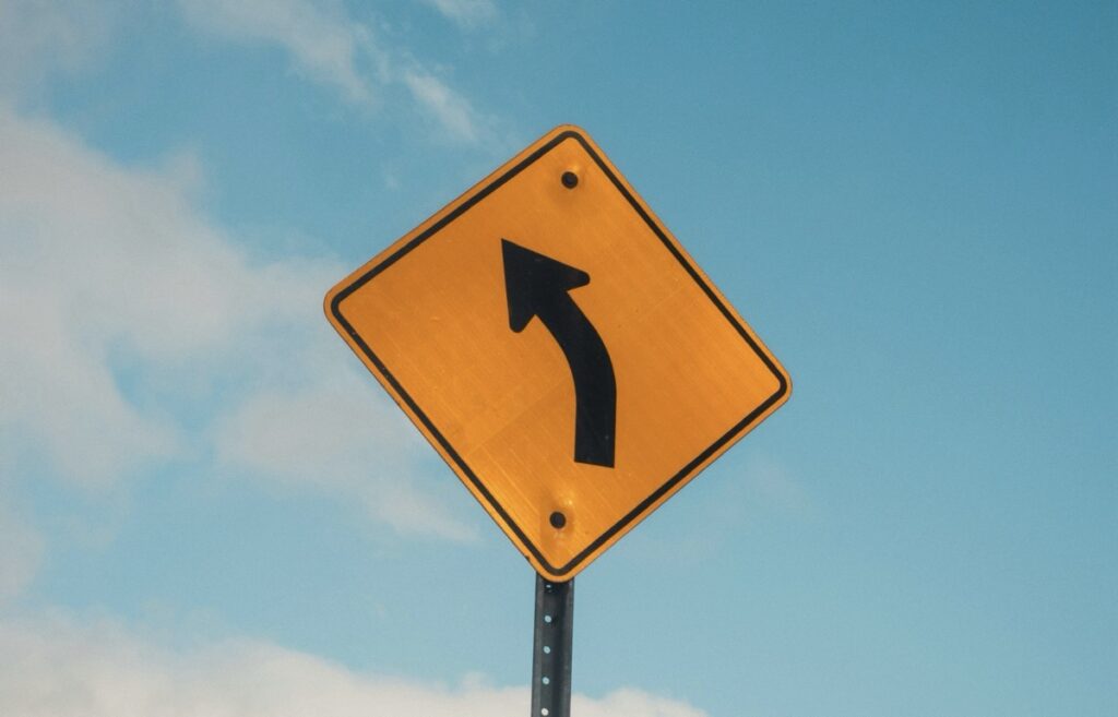A yellow road sign with a black arrow pointing slightly to the left.